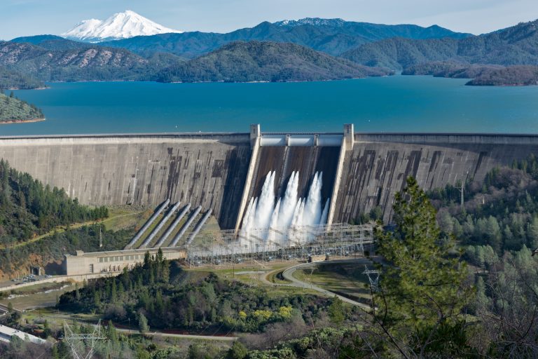 A picture of Shasta Dam surrounded by roads and trees with a lake and mountains on the background