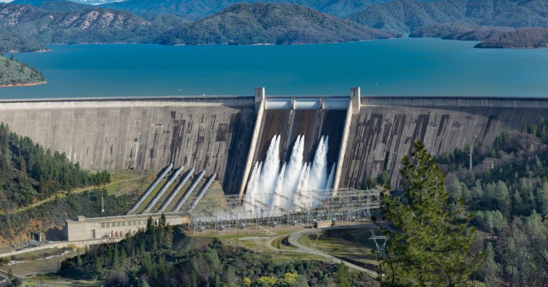 A picture of Shasta Dam surrounded by roads and trees with a lake and mountains on the background