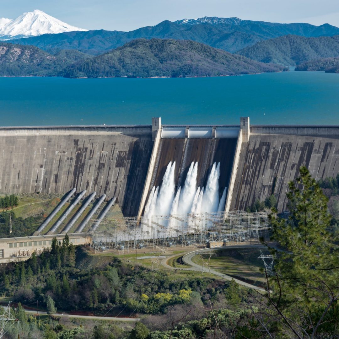 A picture of Shasta Dam surrounded by roads and trees with a lake and mountains on the background
